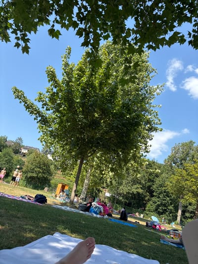 The image depicts a sunny day at a public outdoor swimming area in Bürgstadt, Germany, likely the Main-Badein. The scene is dominated by lush green trees and grass under a clear blue sky with scattered white clouds. Several individuals are visible relaxing on the grassy bank, spread out on towels and blankets. In the foreground, a person's bare foot is visible on a white blanket. Further back, several people are seen interacting, some appearing to play volleyball or another ball game in swimsuits. A small wooden changing cabin is situated on the grass, and a few trees provide shade. The overall atmosphere suggests a recreational and leisurely gathering on a warm day.