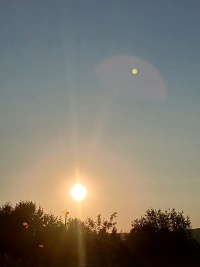The image depicts a rural landscape at dusk in Bolshaya Usa, Russia. The sky transitions from a light blue at the top to a warm orange and yellow near the horizon, suggesting sunset. The sun is a bright, white orb low in the sky, casting a strong glow. Above and to the right of the sun, a smaller, circular object with a textured surface appears in the sky, potentially the moon or another atmospheric phenomenon. Silhouetted against the bright sky are the dark shapes of trees and foliage along the horizon line. In the foreground, a tall object with a distinct spherical or flower-like top is visible, possibly a decorative garden light or a lamp post, with another smaller, red object to its left. The overall scene is serene and captures a moment of natural beauty.
