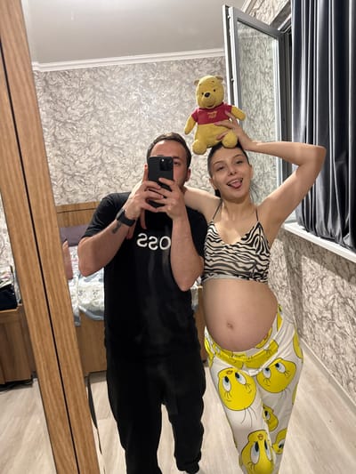 A man and a pregnant woman are taking a mirror selfie in a room that appears to be a bedroom. The man is on the left, wearing a black t-shirt and black pants. He is holding a smartphone to take the picture. The woman is on the right, wearing a zebra-print crop top and white pants with Tweety Bird graphics. She has a Winnie the Pooh stuffed animal balanced on her head and is sticking her tongue out at the camera. The room has wallpaper with a textured pattern, a wooden floor, a bed visible in the background, and a large window with grey curtains. The image was likely taken in Tbilisi, Georgia, given the prompt.