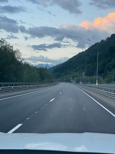 The image captures a view from inside a vehicle driving on a highway in Chvizhepse, Russia. The highway, with its lanes marked by white lines, curves to the right and is bordered by metal guardrails. On the left, a dense forest of trees lines the road, and on the right, a steep, forested mountainside rises dramatically. In the distance, snow-capped mountains are visible against a sky filled with dramatic clouds, some tinged with pink from the setting sun. Several lampposts line the highway, some with what appear to be wind-powered turbines attached. The overall scene suggests a scenic drive through a mountainous region during twilight.