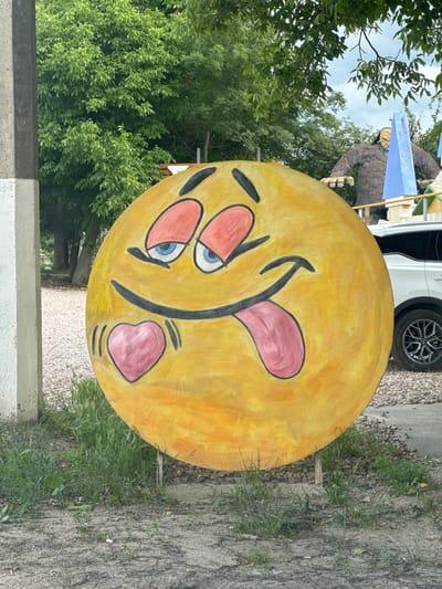 A large, yellow, circular object painted to resemble a smiley face with its tongue sticking out is displayed outdoors. The smiley face has blue eyes, pink rosy cheeks, and a pink tongue. It is positioned vertically in the frame. Behind the smiley face, a large, furry, ape-like statue is visible, along with parts of a white vehicle and a blue triangular flag. Green trees and foliage form the background, suggesting an outdoor setting with a natural environment. The ground appears to be a mix of gravel and dirt with some patches of grass. The lighting suggests it is daytime. The location is identified as Dobrivske settlement, Ukraine. No visible text is present in the image.
