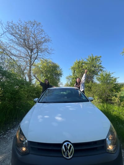 Two people are leaning out of the sunroof of a white Volkswagen automobile. The vehicle is parked on a paved road next to green grass, bushes, and trees under a clear blue sky. Based on the vehicle make, model, and scenery, the location is likely Rish, Bulgaria.