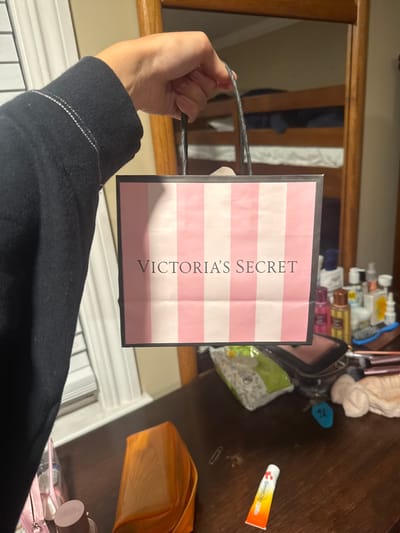 A person's hand and arm, wearing a black hoodie, holds up a small pink and white striped shopping bag with "VICTORIA'S SECRET" printed on it. The bag is the primary subject in the foreground. The background reveals a blurred indoor environment, possibly a bedroom or dorm room, with bunk beds visible. Various objects are scattered on a wooden surface below, including cosmetic bottles, a makeup bag, and what appears to be a tube of ointment. The overall lighting suggests an indoor setting at night or in low light conditions. No specific location in Ocean City, United States of America is depicted.