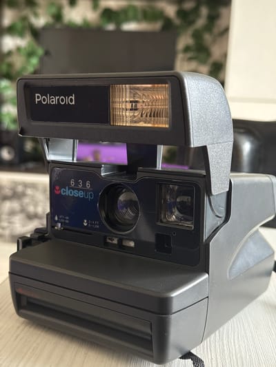 The image shows a Polaroid 636 closeup camera. The camera is positioned on a light-colored, possibly wooden surface. The flash housing is visible on the left, with the "Polaroid" logo prominently displayed. The lens area has a blue sticker with "636 closeup" printed on it, along with distance measurements in feet and meters. In the background, there's greenery suggestive of an indoor plant or outdoor environment visible through a window. There are no people in the frame. The location of Novo-Nikol'skoe, Russia, is not discernible from the image.