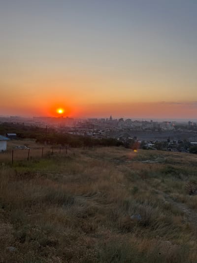 A wide shot captures a sunset over the city of Kok Jar, Kyrgyzstan. The sky transitions from a pale blue at the top to a vibrant orange and red near the horizon, where the sun appears as a bright, circular orb. A hazy cityscape stretches across the middle ground, dotted with numerous buildings of varying heights, some under construction, indicated by cranes. The foreground is dominated by a grassy, undulating hillside covered in dry, golden-brown grass. A rusted metal fence runs horizontally across the hill, and a small, dilapidated white structure with a blue roof is visible on the left. The overall scene depicts an urban landscape at dusk, viewed from a slightly elevated, natural vantage point.