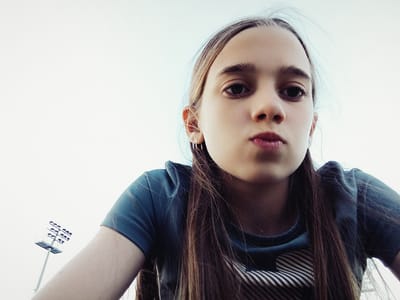 A young girl with long, brown hair is captured from a low-angle perspective in an outdoor setting, likely during daylight given the bright, washed-out sky. She is wearing a teal t-shirt with a striped design on the lower portion of the shirt visible. Her gaze is directed towards the camera, and her lips are slightly pursed. Her left ear is visible, adorned with a small hoop earring. In the background, to the left of the frame, a tall light pole with multiple lamps is partially visible against the sky. The overall scene suggests a casual, possibly recreational environment. The location is identified as Radzymin, Poland.