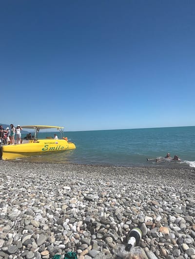 A bright, sunny day at a pebble beach in Amzara, Abkhazia. The turquoise water gently laps at the shore, where a group of people are enjoying themselves. Two individuals are partially submerged in the water, looking towards the camera with smiles. Closer to the shore, a yellow boat named "Smile" is docked. Several people are gathered around the boat, some appearing to be preparing to board or disembark. The boat has a yellow canopy and is emblazoned with the text "Smile J" and a registration number "BE3732RUS23". On the pebble-strewn beach, a bottle of sparkling wine rests near the water's edge, suggesting a relaxed and celebratory atmosphere. The clear blue sky indicates fair weather.