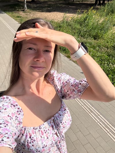 In Kalngale, Latvia, a woman is captured in a selfie taken outdoors on a sunny day. She is wearing a floral-print dress and has her right hand raised to shield her eyes from the sun. On her left wrist, she wears a white smartwatch. The background shows a paved walkway with tactile paving, green vegetation, and trees. The scene suggests a leisurely outdoor activity in a park or along a path.
