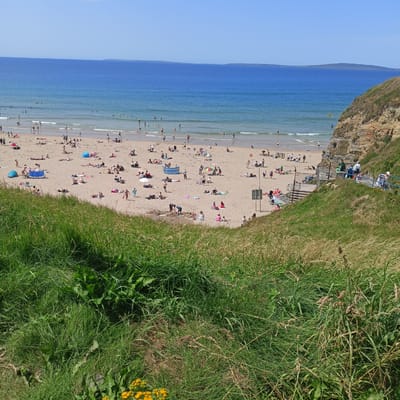 The image depicts a sun-drenched beach scene at Ballybunnion, Ireland, characterized by a wide expanse of sandy shore meeting the blue ocean. The beach is populated with numerous individuals engaged in various activities, including sunbathing on towels and under umbrellas, swimming and wading in the sea, and strolling along the water's edge. Several colorful beach umbrellas and tents are scattered across the sand, adding vibrancy to the scene.
To the right of the beach, a steep, grassy cliff face rises, providing a natural backdrop. A set of concrete stairs, bordered by a blue railing, ascends the cliff, suggesting a path or access point. People can be seen walking up and down these stairs. The foreground of the image is dominated by lush, green grass and vegetation, including some yellow wildflowers, indicating the cliff's natural environment.
In the distance, the ocean stretches towards the horizon, with a faint landmass visible on the far side. The sky is clear and blue, typical of a pleasant summer day. The overall impression is one of a busy and popular recreational area enjoying favorable weather conditions.