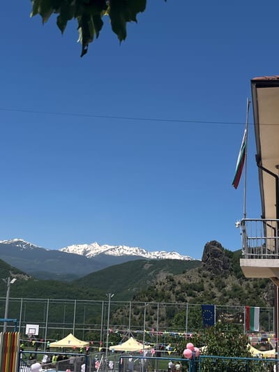 The image depicts a scene in Bukovo, Bulgaria, featuring a building with a Bulgarian flag and European Union flag hanging from it. A mountainous landscape serves as the backdrop, with snow-capped peaks visible. A sports facility, possibly a basketball court, is present with tents and decorative flags. No people are clearly visible.