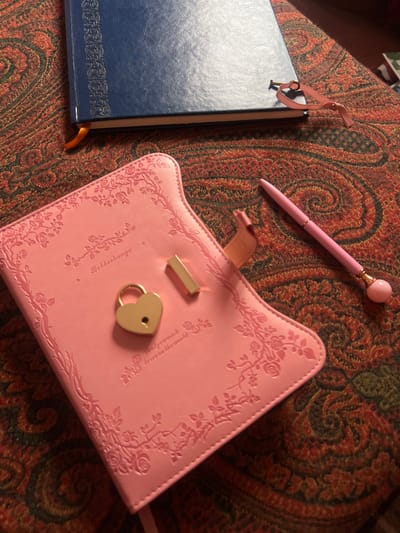 This scene is located in Ksar-el-Kebir, Morocco. The image displays three stationary objects on a patterned textile surface. In the foreground, a pink faux-leather journal or diary, embossed with rose vine patterns and text reading "Betterchange" and "the biggest wish is love in the world," is positioned diagonally. On its surface rests a detached gold-colored heart-shaped padlock with a keyhole, alongside a gold-colored rectangular metal bar, indicating components of a locking mechanism. A pink ribbon bookmark extends from the journal's right edge. To the right of the journal, a pink pen with a gold-colored band and a spherical pink end is angled towards the upper right. Partially visible behind the pink journal is a blue hard-cover notebook or book, featuring embossed designs and a pink ribbon bookmark, with an additional orange bookmark at its lower edge. All items are situated on a fabric surface characterized by a prominent paisley pattern in shades of red, orange, green, and dark brown. No individuals or active interactions are present.