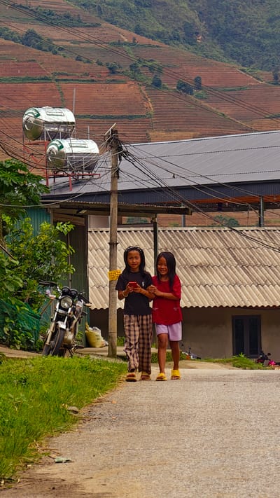 This portrait-oriented image, taken in Pung Luong, Vietnam, displays two young girls walking arm-in-arm on a paved road, set against a backdrop of terraced hills.

The girl on the left, with dark skin and dark hair styled with braids and loose strands, wears a black short-sleeved t-shirt and brown and beige plaid full-length pants. She wears brown open-toed sandals. She holds a red smartphone in her left hand and looks towards the viewer with a slight smile. Her right arm is linked with the left arm of the adjacent girl.

The girl on the right, also with dark skin and long dark hair, wears a red short-sleeved t-shirt and light purple shorts. She wears yellow open-toed sandals and smiles broadly towards the viewer. Her left arm is linked with the first girl's right arm. Both girls are positioned in the center of the frame, walking forward on the light grey paved road.

To the immediate left of the road, a strip of green grass is visible, followed by a dense green bush. Partially obscured by the foliage and a green net is a dark-colored motorcycle, possibly a Honda Wave, featuring chrome handlebars and front fork. Its license plate, "89C1-2401", is partially discernible. A white sack, possibly containing feed or rice, rests on the ground behind the motorcycle.

A prominent utility pole stands to the left of the girls, heavily burdened with numerous black electrical and communication cables crisscrossing overhead. A yellow sticker displaying "0312" is affixed to the pole. Above and behind the pole, two large, shiny, cylindrical metallic tanks, likely solar water heaters, are mounted on a rooftop structure. One tank clearly bears the branding "BIOTRAMIX".

Behind the utility pole and tanks, a building with a light grey corrugated roof and light-colored walls is visible, featuring a dark rectangular window on its lower right. Further back, another structure with a dark metal roof can be seen. In the background, steep mountains rise, characterized by extensive reddish-brown agricultural terraces, interspersed with patches of green trees and vegetation. The scene is illuminated by an overcast or diffused light.