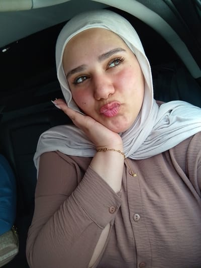 A woman is captured in a close-up selfie taken inside a car, likely as a passenger. She is wearing a light-colored (off-white or light grey) hijab that drapes around her head and neck, and a long-sleeved, collared shirt in a muted brown or taupe color with a textured fabric. Her right hand is raised, with her chin resting on her palm, and her lips are pursed in a "kiss" face. Her eyes are looking upwards and slightly to the right, away from the camera.

Notable details include visible mascara on her long eyelashes, pink blush on her cheeks, and a glossy pink lip color. She wears a delicate gold-colored chain bracelet with a small charm on her right wrist. Her fingernails are long and pointed, with a light-colored polish. The background shows the dark interior of the car, with hints of the ceiling or headliner visible. A fragment of a bag is seen in the bottom left corner, revealing the letters "MK" printed on its pattern, suggesting it might be a Michael Kors bag. The overall mood is playful and a bit flirtatious. The location context provided is Qudsaiya, Syria.