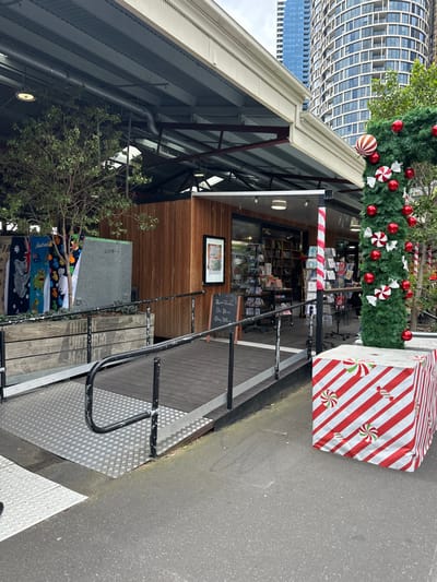 A commercial building with a wood facade is adorned with Christmas decorations, including a garland, ornaments, and a wrapped box. A black metal railing is present. The building's interior reveals rows of products, possibly books or cards. A frame hangs on the exterior wood panel. A grey structure and foliage are visible on the left, and the building is framed by taller structures and the sky.