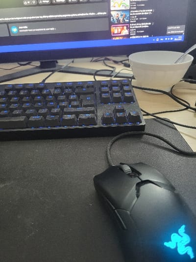 A black keyboard with blue backlit keys sits on a black mouse pad. A black gaming mouse with a green and blue light on the bottom sits on the pad. There is a white bowl on a wooden desk to the right of the keyboard. The bowl is empty. There is a black cable connected to the mouse. The image was taken in Louches, France, located at 50.829 latitude, 1.997 longitude.  The image was taken from an elevated angle, looking down at the desk. The background is blurred.