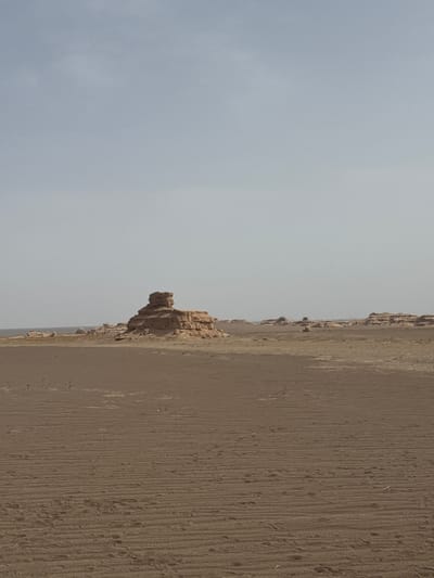 The image depicts a landscape in Dunhuang City, China. The scene is dominated by a desert or barren terrain on the right side, characterized by ridges and texture. On the left, a greyish sky occupies a significant portion of the frame.  In the mid-ground, there are scattered rock formations along the horizon line.