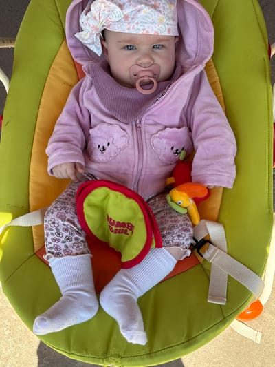 A baby lies in a green and orange baby bouncer, positioned on an outdoor surface. The baby wears a pink hooded jacket with cloud-shaped appliques, floral-patterned pants, and white socks. A pacifier is in the baby's mouth, and a floral-patterned hat adorns their head. A plush toy rests near the baby's side. The baby bouncer has a white strap. The image provides no discernible evidence to confirm the specific location is Gorna Mitropolia, Bulgaria.