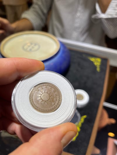 A close-up, shallow depth of field shot shows a person's hand holding a coin in a clear protective case. The coin, which appears to be made of a copper-colored metal, is round and features a sunburst design in the center with Chinese characters surrounding it. The protective case has a white, ridged insert. In the background, out of focus, a person wearing a striped shirt is visible, along with a blue ceramic bowl and other objects on a dark surface. The overall scene suggests a casual display or examination of the coin. The location is not discernible from the image.