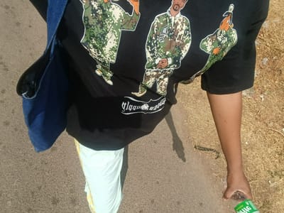 A person is standing on a paved surface, likely a road or sidewalk. The person is wearing a black t-shirt with a graphic print of three cartoon figures in camouflage outfits, a white lower garment with gold accents, and a blue denim bag is slung over their shoulder. They are holding a green and clear plastic bottle in their right hand. The surrounding environment appears to be an arid or dry landscape, with dry grass. The location is Rafin Pa, Nigeria.
