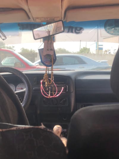 The image depicts a scene observed from within a vehicle in Basawa, Nigeria, specifically from the rear seat looking forward through the windshield on a bright, hazy day.

The foreground shows the interior of a vehicle. A dark-colored dashboard occupies the lower half. On the left, a portion of a dark steering wheel with a patterned cover is visible, revealing a white-faced instrument cluster with red needles. The center console contains air vents, climate control knobs, and a car stereo unit displaying a red indicator light. A light-colored gear shifter knob is positioned between the front seats. Hanging from the rearview mirror are a blue and white decorative card, a set of brown beaded prayer beads, and a pink coiled cable looped below them. The rearview mirror itself is slightly angled. The windshield exhibits multiple cracks, particularly in the upper and central areas, and features a blue sun strip along its top edge. A dark headrest is partially visible in the lower right, and a patterned fabric cover is seen on the lower left seat.

Through the windshield, two other vehicles are visible: a red car partially obscured on the left, and a silver or white sedan with a rear spoiler positioned further to the right. The background features light-colored buildings under a bright sky, with a partial blue and white circular logo or sign visible on the far right. Sparse vegetation is also present. The overall environment suggests an outdoor setting during daylight hours, possibly with some atmospheric haze.