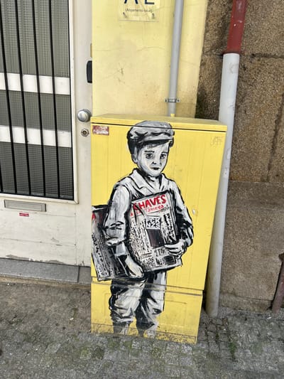 A street scene in Chaves, Portugal, features a grayscale mural of a young boy painted on a weathered yellow utility box. The boy, depicted wearing a cap and jacket, holds multiple newspapers under his left arm and prominently displays one in his hands. The visible headlines on this newspaper include "CHAVES" and "TÂMEGA" in red capital letters, with "Notícias" also discernible.
The utility box stands on a grey cobblestone sidewalk. To its immediate left, a distressed white wooden door with a rectangular, gridded window section is visible, featuring a silver doorknob and a mail slot. A grey concrete threshold extends from the base of the door. Above the utility box and door, a pale yellow stucco wall exhibits minor cracking and holds a clear acrylic sign with black text, partially reading "AL (Alojamento local)". To the right of the utility box, a rough-textured stone wall is present. A grey utility conduit runs vertically down the yellow wall, and a red-capped white pipe along with a narrower, rusty red pipe emerge from the ground against the stone wall. The foreground consists of weathered, uneven grey cobblestones.