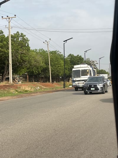 The image, taken from within a vehicle, depicts a road in Galadimawa, Nigeria. A black SUV is traveling on the right side of the road, followed by a white truck loaded with various objects. The road is paved and appears to be in fair condition. On the left side of the road are trees and utility poles. The sky is overcast. No people are clearly visible.