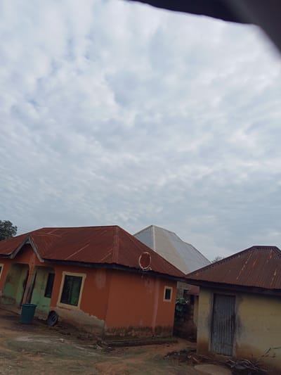 The image depicts a collection of modest buildings with corrugated metal roofs under a cloudy sky. The buildings are situated in what appears to be a rural or semi-urban setting in Gindin Dutse, Nigeria. The primary focus is on the structures themselves, which vary in color and condition. One prominent building has an orange-red facade and a rusty corrugated iron roof. To its left, another structure with a greenish-blue facade and a similar roof is visible. A dark green trash can and a spare tire are on the ground in front of these buildings. To the right of the orange-red building, a smaller structure with a light-colored, weathered facade and a corrugated iron roof is present. A dark, weathered door is visible on this smaller building. In the background, a larger building with a white, angular, metallic roof stands out. The ground appears to be dirt and gravel, with some vegetation and debris scattered around. The overall impression is one of a functional, lived-in environment with utilitarian structures.