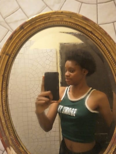 A female subject is depicted in a mirror selfie taken indoors. She has dark, medium-length, voluminous hair and wears a dark green crop top with white trim and "BROOKLYN" in white text (reversed), exposing her midriff. Small dark earrings are visible. She holds a dark-colored smartphone in her left hand, positioning it to capture her reflection in an oval mirror. The mirror features an ornate golden frame with visible signs of age and wear. The reflection reveals a pale yellow wall behind the subject and a dark rectangular vertical element, possibly a door or doorway, to the reflected left. To the reflected left of the subject, and visible beyond the mirror's frame, is a wall tiled with light-colored square tiles configured in a radial or concentric pattern. The specific location within Cotonou, Benin, cannot be determined from the visual information provided.