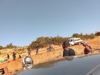 The image depicts an outdoor scene in Ibi, Nigeria, characterized by dry, reddish-brown earth and a clear blue sky. The ground slopes upwards from left to right, featuring scattered green bushes and small trees. A dirt path or road traverses the midground horizontally.
In the immediate foreground, the dark gray roof and side of a vehicle are partially visible, with a hand in the lower-left corner. From this vehicle, a male with dark skin and short hair, wearing a black t-shirt with a white shoulder stripe, is visible from behind, facing right. Next to him, a female with dark skin, wearing a red top and a patterned red, white, and dark headscarf, is visible in profile, also facing right. Partially obscured below them is an individual with a white, orange, and black patterned head covering.
On the dirt path and sloping terrain, several individuals are present. Towards the far left, a person in a white top and dark shorts stands near a light blue basin. Further right, a man in a yellow shirt stands, and another in a dark top with a red detail is visible. A man in a blue shirt and dark shorts stands further right. A dark-colored motorcycle with a rider is centrally positioned on the path, and behind it, a man in a blue hoodie is visible. In the upper-right midground, a white flatbed truck loaded with multiple light-colored sacks is parked on the dirt road, adjacent to a vegetated hillside. Stacked wooden planks or construction materials are on the ground to the right of the central foreground figures.