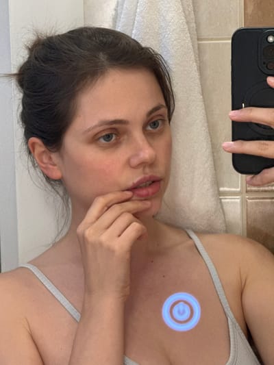 A woman takes a selfie in a bathroom. She holds a black smartphone in her left hand, partially obscuring her face. She wears a grey tank top. Her hair is dark brown, and her eyes are blue. The bathroom features light-colored tiles and a white towel. A blue concentric circle graphic is superimposed on her chest. There is no information available to confirm whether this photo was taken in Kőröshegy, Hungary.
