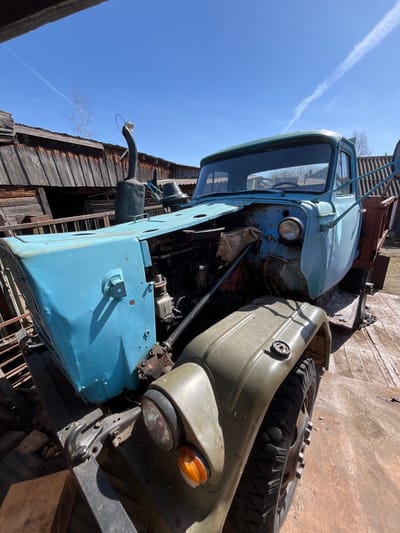 The image shows a dilapidated, light blue and olive green truck with missing body panels. The truck is situated in what appears to be an outdoor storage or salvage yard, with an old wooden building visible in the background. The sky is clear with a contrail. I do not have information about the specific geographic location of the photo.