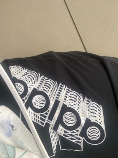 The image shows a close-up of a black t-shirt with a white Adidas logo printed on it. The t-shirt is laid flat, and part of another garment with a floral pattern is visible underneath it. A white hanger is also visible, positioned diagonally across the t-shirt, obscuring part of the logo. The background appears to be a light-colored, textured surface, possibly a wall or floor. The lighting is somewhat dim, creating shadows on the t-shirt. The scene is photographed from a slightly elevated angle, looking down at the garments. There are no people visible in the image. The location of Wauro Leggal, Nigeria is not evident from the visual information provided in the image.