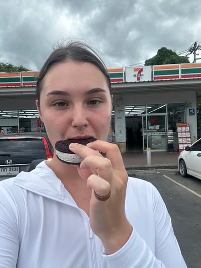 A woman in a white zip-up jacket holds an ice cream sandwich to her mouth, as if to take a bite. She is in the foreground of the image, and behind her is a 7-Eleven convenience store. The store is a typical urban convenience store with glass doors and windows, and a red, orange, and green striped sign with "7 ELEVEN" in white lettering. A white car is parked to the right of the store, and a black SUV is to the left. License plates on both vehicles are visible and appear to be Thai. The ground in front of the store is paved with asphalt. The sky is overcast and appears to be cloudy, suggesting it may be raining or about to rain. The overall scene is one of a casual outdoor setting in a commercial area.