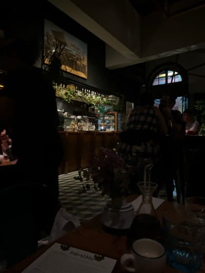 The image shows a dimly lit indoor space, likely a restaurant or bar, in Barranco, Peru. A person wearing a plaid shirt stands near a table with a vase of flowers, a glass of water, a mug, and a menu. In the background, there is a bar stocked with bottles and decorated with greenery. A painting hangs on the wall. A round window is visible. People are gathered in the background, some standing near the bar.