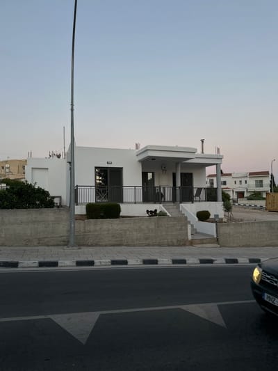 The image depicts a street scene in Peyia, Cyprus, at dusk. A modern, single-story white house with dark railings and a covered porch is the central focus, situated behind a concrete retaining wall and a sidewalk. The house has large glass sliding doors and windows, reflecting the sky and surroundings. To the left of the house, a dense green bush is visible. A tall, grey lamppost stands prominently in the foreground, slightly to the left of center. Across the street, a dark-colored car with its headlights on is partially visible on the right side of the frame, driving on an asphalt road marked with a white directional arrow. In the background, other houses with tiled roofs can be seen, along with some construction elements, such as rebar protruding from a building on the left. The sky is a pale gradient of blue and pink, indicative of twilight.