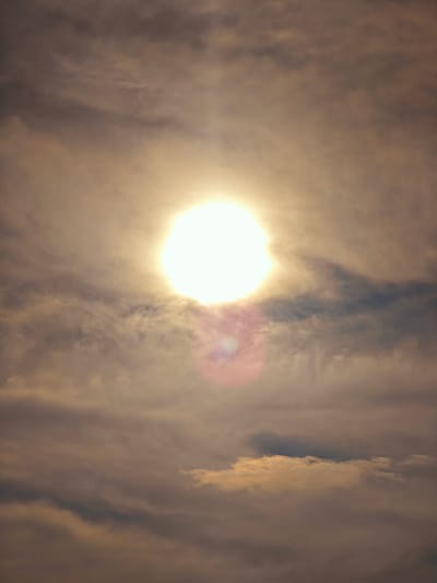 A high-angle, close-up shot captures the sun in the sky, partially obscured by clouds. The sun is a bright, glowing orb in the center of the frame, emitting a strong glare. Wispy, grey and tan clouds swirl around the sun, creating a textured and atmospheric effect. The overall lighting is soft and diffused, suggesting either early morning or late afternoon. The scene is likely outdoors and depicts a natural sky formation. No specific landmarks or structures are visible to identify a precise location within Юксеево, Russia.