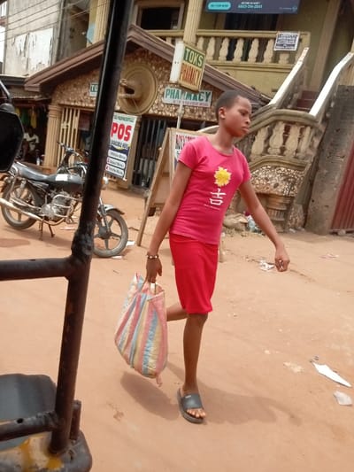 The image captures a young female walking on an unpaved, reddish-brown dirt road in Umudioka, Nigeria. She is attired in a short-sleeved red t-shirt featuring a yellow floral design and a Chinese character, a red knee-length skirt, and dark grey slides. A multicolored striped woven bag is carried in her left hand, which also displays a light-colored bracelet. Her head is turned slightly to her right. In the background, a two-story building with a decorated facade and a brown roof is visible. Signage on the ground floor identifies "TOKS PHARMACY" and advertises "POS AVAILABLE HERE" services. An "MTN HYPE NOW" banner with a phone number is affixed to the upper level of the building. A dark-colored motorcycle is parked on its stand to the left of the building. A dark, metallic frame structure, possibly part of a vehicle, partially obstructs the lower-left foreground. The scene is illuminated by bright daylight, casting short shadows.