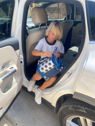 A young Caucasian child with blonde hair is sitting in the open rear door of a white car. The child is wearing a white t-shirt with a blue collar and a logo on the left chest, and shorts with a soccer ball pattern. The child is holding a blue backpack adorned with various sports motifs, including basketballs and soccer balls. The backpack's zipper is partially open, and the child's hands are near it. The child's feet are clad in white sneakers with white laces. The car's interior features beige leather seats. The scene is set outdoors, with a paved surface visible beneath the car and a wooden fence in the background, suggesting a residential or private area. The lighting indicates it is daytime, likely sunny. The location is identified as the city of Bel Air, Spain.