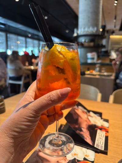 A close-up view from within a modern restaurant, identified as likely Sushi Suzan in Kiryat Ono, Israel, based on visible menu branding. A left human hand, featuring light pink manicured nails and a gold-toned ring on the ring finger, holds a stemmed glass. The glass contains an orange-colored, effervescent cocktail with ice and orange slices, fitted with two black straws. The hand and glass are positioned above a light-colored wooden table. Two black-covered menus, one displaying "SU SU" text and a barcode, rest on the table. The blurred background reveals additional restaurant patrons seated at tables, large windows with horizontal blinds, a dark ceiling, and a concrete cylindrical pillar.