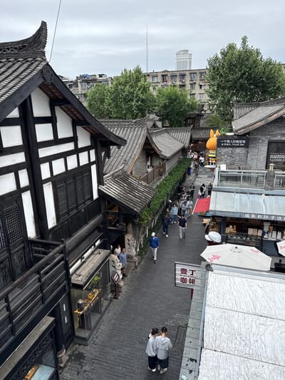 This elevated, overcast daytime image depicts a bustling pedestrian street in Qingyang District, China, showcasing a blend of traditional and modern urban landscapes. The narrow street, paved with dark grey interlocking bricks, is bordered by traditional Chinese-style buildings featuring dark timber frames, white walls, and intricate dark tiled roofs with upturned eaves. Windows are detailed with dark wooden grilles.

Numerous pedestrians, dressed in casual contemporary attire, are seen walking along the thoroughfare. On the right, a large yellow anthropomorphic figure stands beside a black sign for '一个男人的咖啡馆 a man, must be'. Further along, a shop with a grey awning displays Chinese text. On the left, a traditional building features a ground-level glass display case exhibiting what appear to be cultural artifacts or tea sets. Balconies with dark wooden railings are visible on these traditional structures.

In the foreground, two individuals stand close together. A man in a blue jacket and white cap is walking toward the foreground on the left. A white umbrella with pink Chinese characters and a 'Ding's Coffee' sign are visible on a nearby rooftop terrace, part of which is covered with a silvery, textured insulating material.

The background reveals a modern urban environment with dense green trees, concrete apartment buildings featuring numerous air conditioning units, and a distinctive tall, white high-rise building with a curved top. A slender communication tower is also discernible against the grey sky.