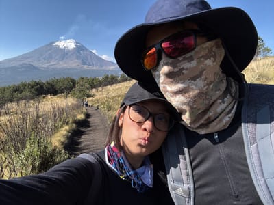 A selfie shows two people on a mountain trail, possibly at the location of Tlamacas, Mexico, given the presence of an unnamed mountain. The person in the foreground wears sunglasses and a hat, with the lower face covered by patterned fabric. A second person is behind, wearing glasses and a patterned headband. The background includes a barren trail, sparse trees, and a large mountain with snow on the summit. The sky is blue with minimal clouds.