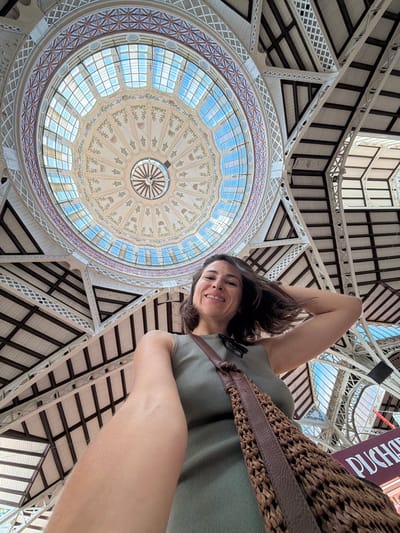 An indoor, low-angle, wide shot captures a woman taking a selfie in what appears to be a large public market. The woman, in the foreground, is wearing a sleeveless, olive-green ribbed tank top and has a brown woven shoulder bag with leather straps slung across her chest. Her right arm is raised, touching her hair, and she smiles directly at the camera. The background is dominated by an ornate domed ceiling with stained glass windows allowing natural light to filter through. Intricate decorative patterns adorn the dome, featuring floral motifs and geometric designs. The structure is supported by a complex network of white metal beams and dark wooden rafters. A sign with "PUCHAR" is partially visible in the bottom right corner. This setting is the Mercado Central in Valencia, Spain, a prominent architectural landmark.