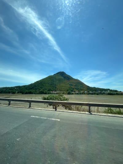 A wide-angle, full shot captures a lush green mountain with a communications tower on its summit, situated in the city of Tuy Hoa, Vietnam. The mountain, covered in dense vegetation, slopes down towards a flat expanse of agricultural fields, likely rice paddies, that appear to have been recently harvested or are awaiting planting. A light brown metal guardrail runs horizontally across the middle ground, separating the road from the fields. In the foreground, a paved road, possibly a highway, is visible, with a single white line marking the lane. The sky above is a vibrant blue, dotted with wispy white clouds that stretch across the frame, suggesting a sunny day. The overall scene depicts a rural or semi-rural landscape on the outskirts of Tuy Hoa, characterized by the juxtaposition of natural topography and human cultivation.