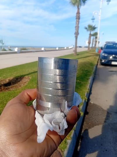 A hand holds a cylindrical object with vertical markings wrapped in a white tissue. The background includes a paved area, grass, palm trees, streetlights, a dark-colored car, and a blue sky with clouds. The scene appears to be located in Martil, Morocco.