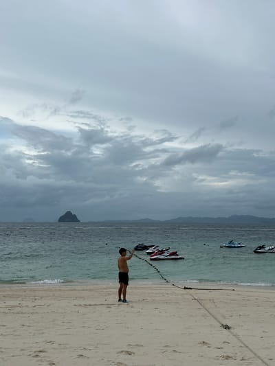 A man stands on a sandy beach in Tambon Phru Nai, Thailand, holding a rope that extends into the water. The sky is overcast with gray clouds. The ocean is a muted blue-green, with gentle waves lapping at the shore. Several jet skis are visible in the water, some clustered together and others more spread out. In the distance, a rocky island with a pointed peak rises from the sea, and a hazy mountain range forms the horizon. The beach is made of light-colored sand with some footprints visible. A thick rope is anchored to the sand and stretches out into the water, possibly to moor the jet skis.