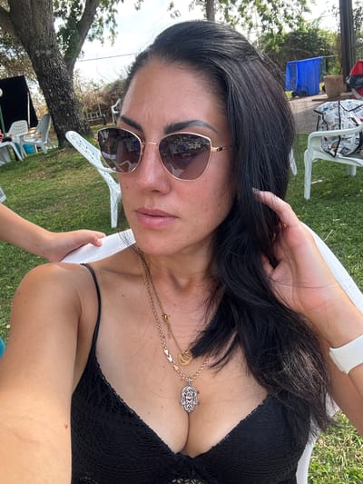 A woman with long, dark hair and sunglasses is seated outdoors. She is wearing a black bikini top and multiple necklaces. Her left arm is resting on the back of a white chair, and her right hand is near her face, touching her hair. Behind her, a child's arm is visible resting on her shoulder. The background consists of a grassy area, trees, and some outdoor furniture. The scene appears to be a backyard or park setting in Beit Yitzhak-Sha'ar Hefer, Israel.
