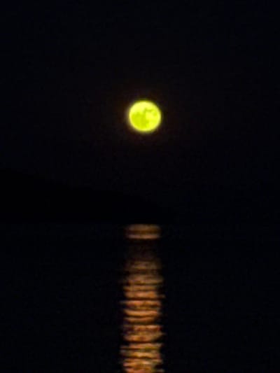 A full moon, colored a vivid yellow, hangs in the center of a black sky. Below the moon, a reflection of the moon shimmers on a dark body of water, appearing as a blurry, vertical column of orange and yellow light. The scene is devoid of any other visible elements, suggesting a tranquil, nocturnal environment. This image was captured in the Municipal Unit of Tolofona, Greece, a location known for its natural beauty and clear night skies.