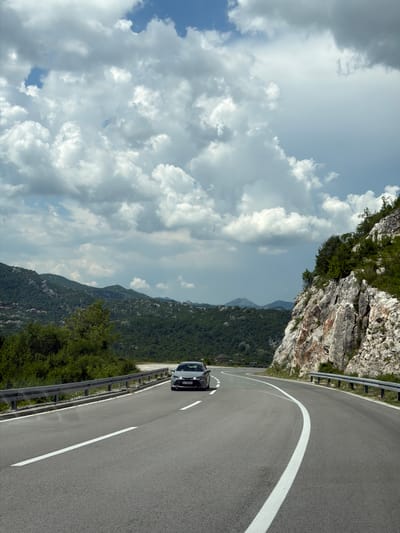 A dark sedan travels on a winding asphalt road with white lane markings. The road is bordered by a metal guardrail on the left and a rocky, vegetated hillside on the right. In the background, lush green mountains rise under a cloudy sky. The scene appears to be a scenic route in Obzovica, Montenegro.