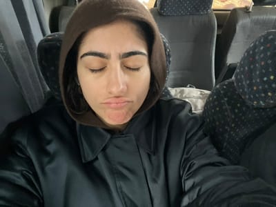 The scene depicts the interior of a vehicle, likely a bus or large van, in the city of Zerkten, Morocco. A single female individual is centrally positioned, seated with her eyes closed and her mouth pursed into a pout, causing a slight wrinkle in her chin. She possesses dark, prominent eyebrows and is wearing a dark brown hooded garment over a dark, collared jacket or coat with a subtle sheen. The surrounding environment consists of multiple rows of grey upholstered seats; those to the right feature a distinct pattern of small, multi-colored dots. A dark, vertical fabric element, possibly a window curtain, is visible on the left side of the frame. The lighting within the vehicle appears subdued.
