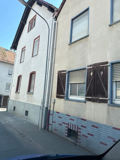 This image captures an exterior view of buildings with a focus on architectural details. The scene appears to be a residential or commercial area in Weidenhausen, Germany. The buildings have textured off-white facades and various window designs, including some with brown wooden shutters featuring diamond patterns. There are also modern windows with slatted coverings.
The composition is characterized by an angled perspective, possibly taken from a car or an elevated position, looking up at the buildings. A prominent feature is the brickwork on one side of a building, a mix of grey and reddish-brown bricks arranged in a pattern. Plumbing and drainage pipes are visible along the exterior walls, indicating functional elements of the structures.
The lighting suggests it is daytime, with clear skies visible at the top of the frame. The overall impression is one of everyday urban or suburban architecture. No people or specific activities are depicted. The image does not contain any visible text.